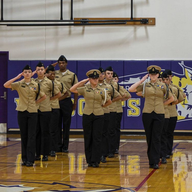 A group of young people in military uniforms stand in formation, saluting in a gymnasium.