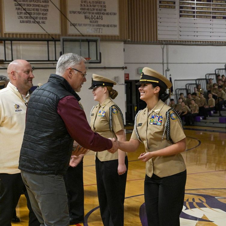 A man in a maroon shirt and black vest shakes hands with a woman in a navy uniform, smiling.