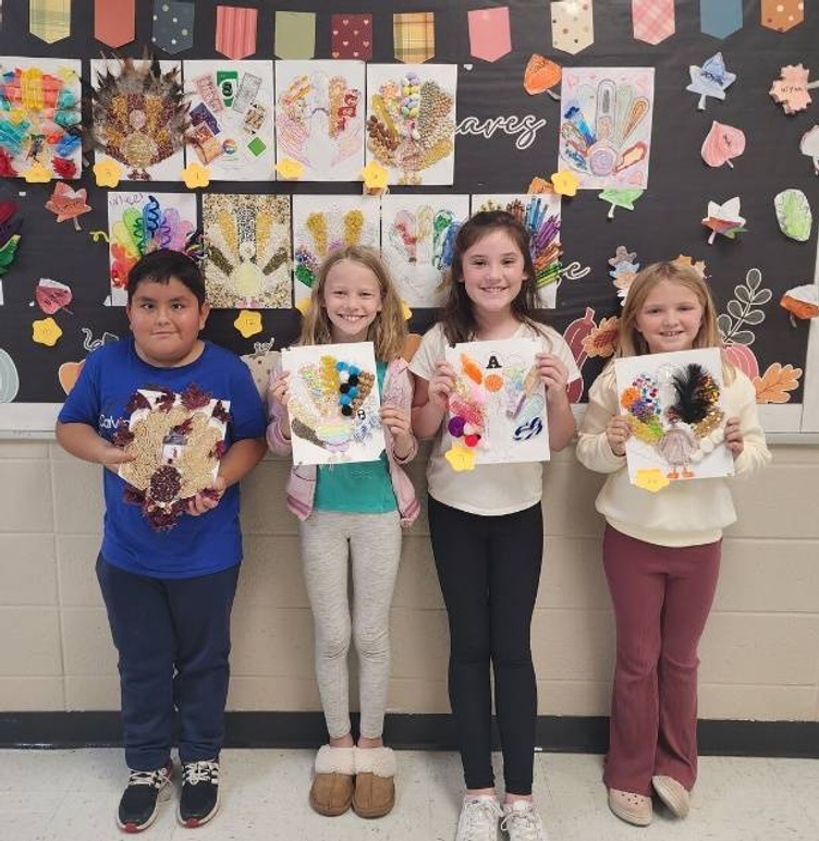 Four elementary students pose with their disguised turkeys, standing before a bulletin board filled with other students' disguised turkeys.