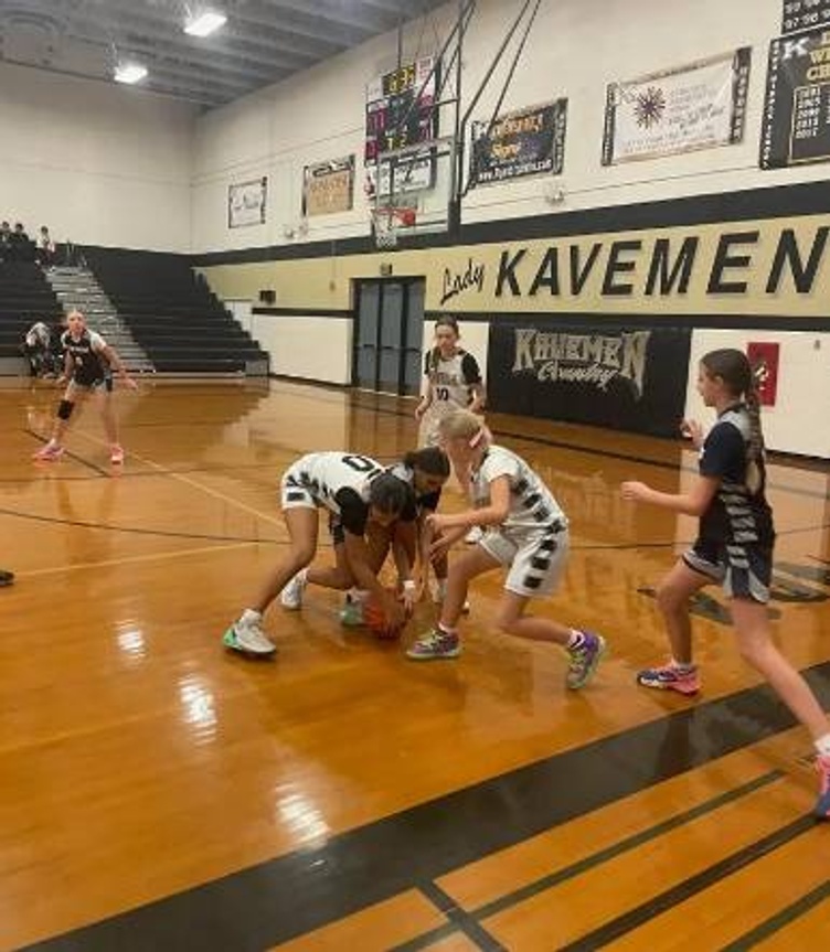 Four young girls compete for the basketball during a game, with one girl in a blue and white uniform running towards the ball.