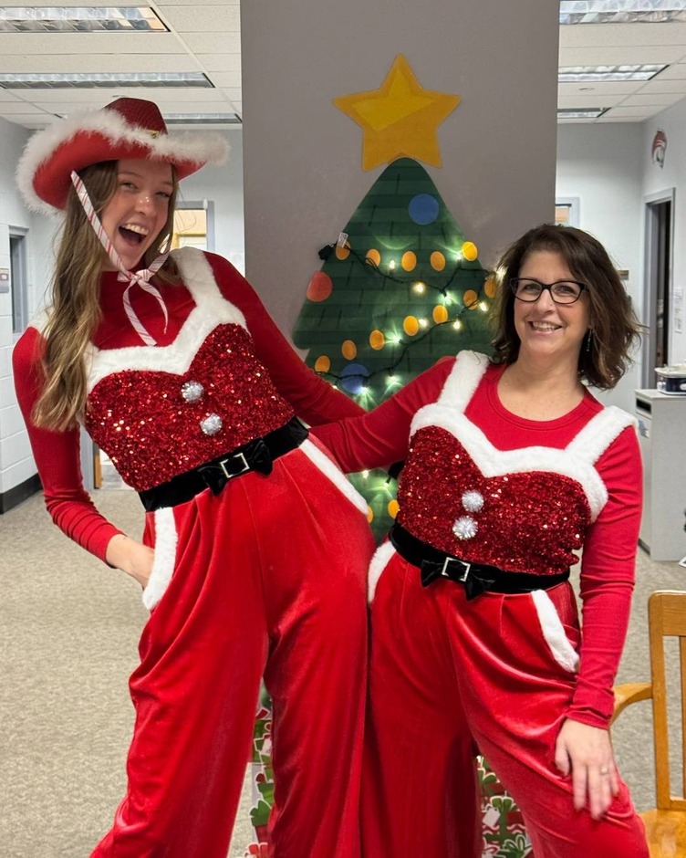 Two women in festive red and white outfits pose in front of a Christmas tree, smiling for the camera.