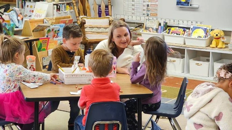 A photo of four transitional kindergarten students sitting at a table in a brightly decorated classroom, with their teacher in a white sweater crouching at their table. The kids are coloring and the teacher is speaking to a girl. 