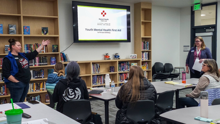 Two women stand in front of a group inside a school library, with a presentation titled "Youth Mental Health First Aid" on a large flat screen up on the wall. 