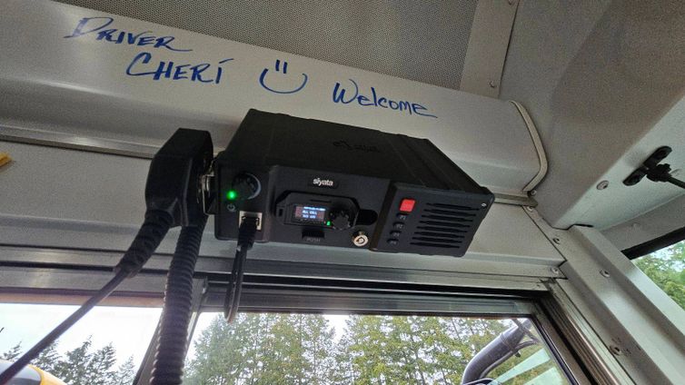 Close-up of a new Siyata radio communication system installed inside a school bus, with a handwritten message above it reading, 'Driver Cheri :) Welcome.' The view outside the bus shows trees in the background