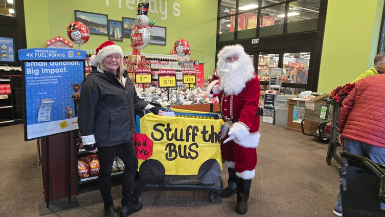 Santa Claus stands next to a shopping cart decorated for a food drive, encouraging donations with the sign "Stuff the Bus."