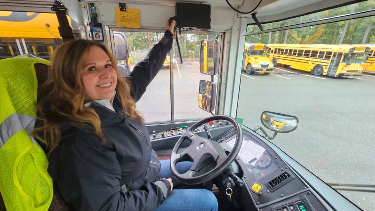 A school bus driver smiling while seated in the driver’s seat of a school bus. She is reaching up to adjust or test a newly installed radio communication system inside the bus. Through the windshield, other yellow school buses are visible in the parking lot.