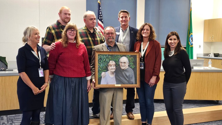 A group of eight adults stands together, smiling, in a formal setting with an American flag and Washington State flag in the background. The man at the center holds a framed portrait of an elderly couple, both smiling warmly in the painting. The group appears to be honoring or celebrating the couple in the portrait, possibly in a ceremony or recognition event. The individuals are dressed in professional attire, creating a respectful and appreciative atmosphere.