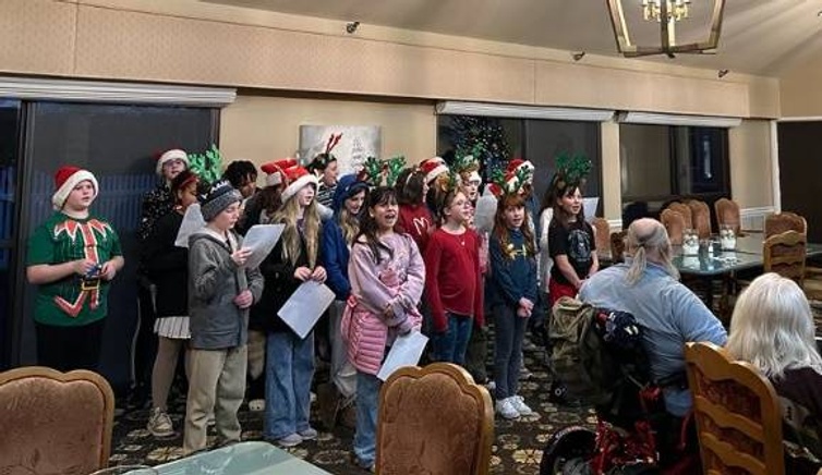 A group of children in festive attire sing carols for a seated audience.