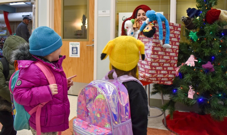 Two children wearing backpacks look at a decorated Christmas tree with a woman standing behind it.