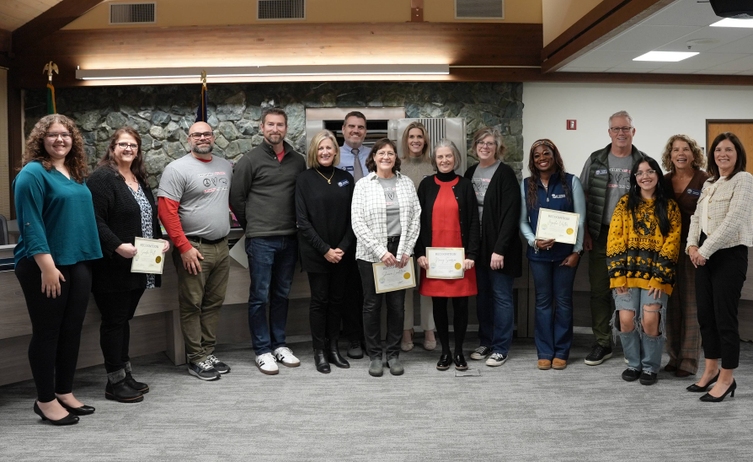 A group of people stand in a room, holding certificates of recognition. The text on the certificates reads 'Recognition' and includes the names of the recipients.