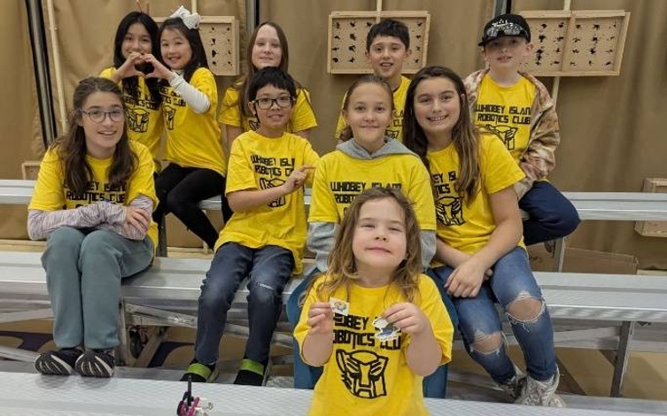 A group of children, wearing yellow shirts with the words 'Whidbey Island Robotics Club', pose for a photo on bleachers.