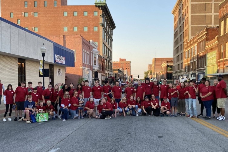 A large group of Peer Buddies students wearing matching maroon tshirts are posed together across two street lanes in the middle of the road before the parade begins.