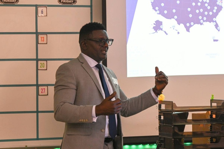 A man in a suit gestures while presenting in front of a map and a calendar.