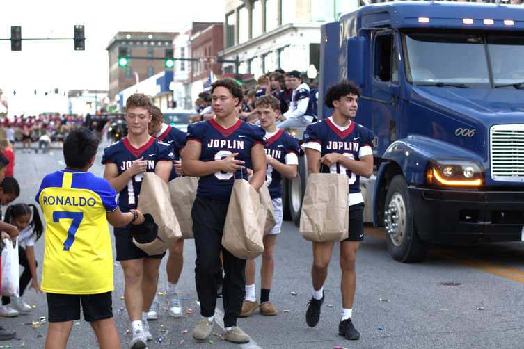 JHS Football players walk beside a flatbed trailer pulled by a semi-truck; the players are wearing their jerseys, interacting with bystanders, and passing out candy.