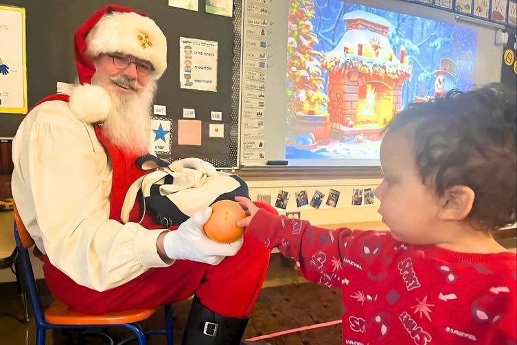 Santa Claus sits on a chair in a classroom, holding an orange while a child reaches out to touch it.