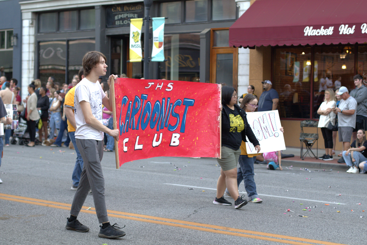 Students walk down the center of main street carrying a red banner declaring "JHS Cartoonist Club" in hand-painted artistic fonts.