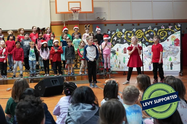 A group of children perform a holiday play on stage, with a festive backdrop and audience members watching.