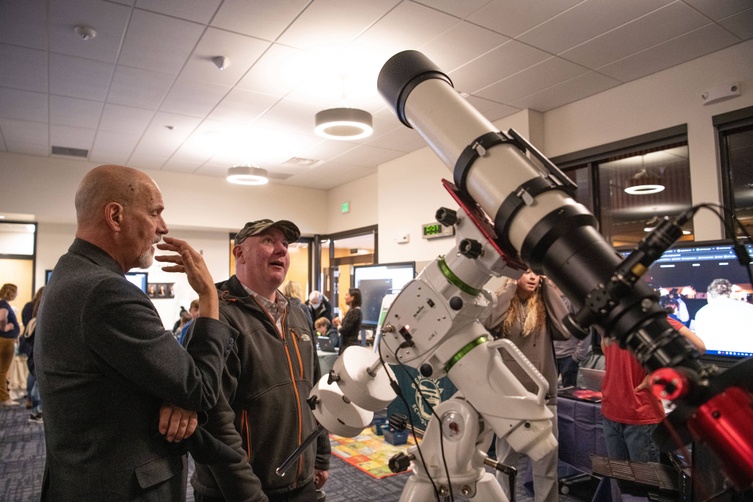 Two men engage in animated conversation next to a large telescope at the Focus on Education Innovation event. They stand in an indoor space with circular ceiling lights, and other attendees can be seen in the background.