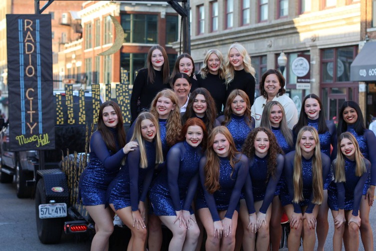 Members and coaches of the JHS Dance team are posed on their Radio City Music Hall-themed float, smiling in matching blue sequined uniforms or black Rockettes costumes.