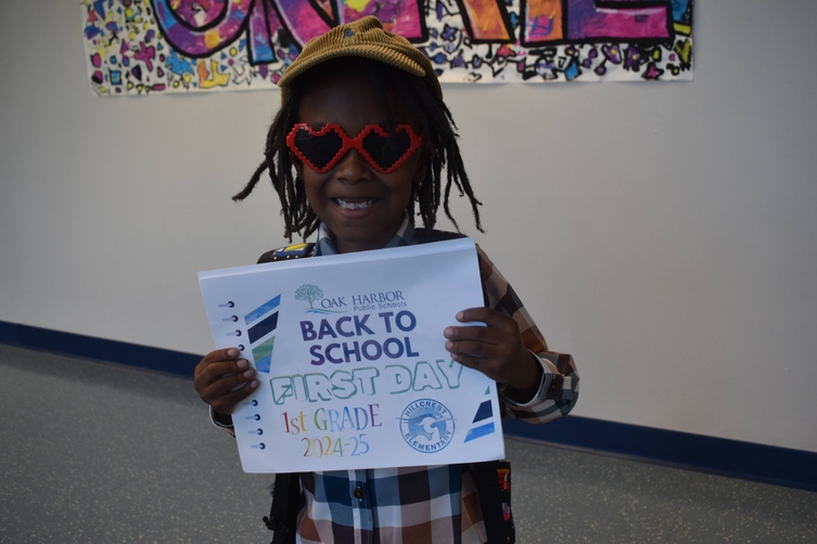 Kindergarten student wearing hear shaped sunglasses hold in sign with text "Back to school first day of first grade 2024-25ts