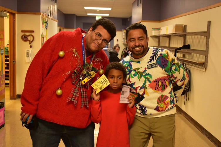 Three people, including a child, stand in a hallway, smiling for a photo. The two adults are wearing festive sweaters.