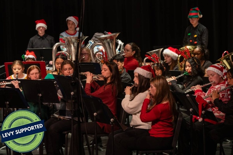 A group of musicians, many wearing Santa hats, play flutes during a holiday performance. The sign reads 'Levy Supported'.