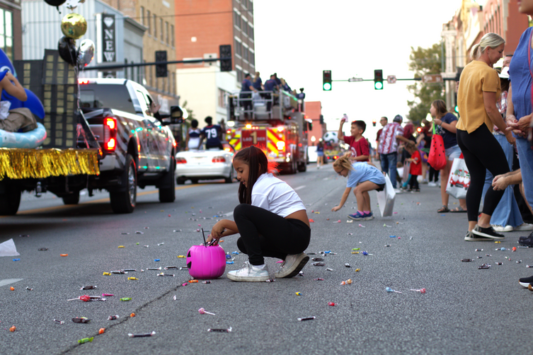 A young girl holding a pink pumpkin bucket kneels down to collect candy scattered on the ground. In the background, more kids scramble for candy as parade floats slowly continue down the center of Main street.