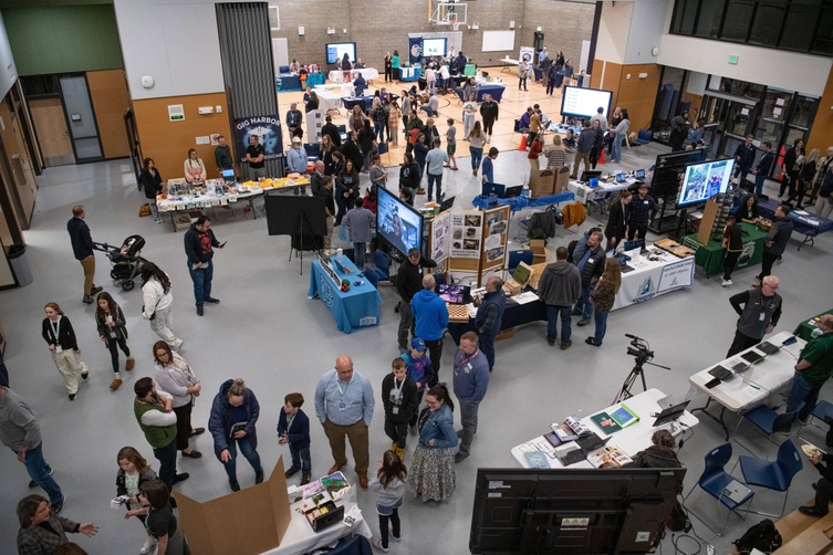 An aerial view of the busy Focus on Education Innovation showcase in the Swift Water Elementary School commons and gym. Multiple tables are set up with displays and banners, while crowds of people move between booths, engaging with presenters and viewing exhibits