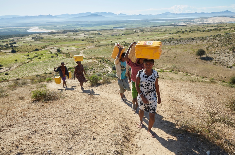Fem människor med vattendunkar går upp för en sandig stig i värmen på Madagaskar.