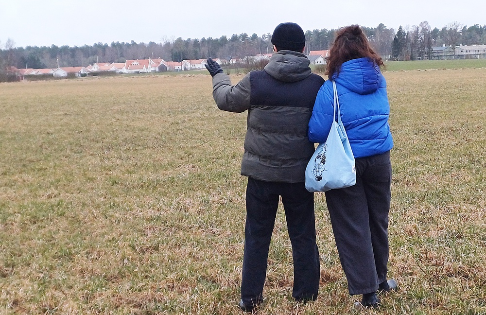 Två unga personer tittar ut över ett obebyggt fält med bebyggelse i bakgrunden.