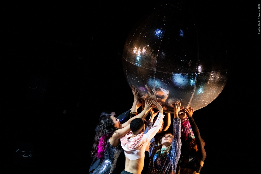 Ett gäng dansare håller en stor silverfärgad discokula över sina huvuden.