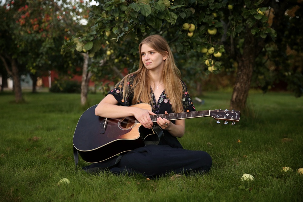 Kvinna i långt hår sitter i gräset med gitarr i knät.