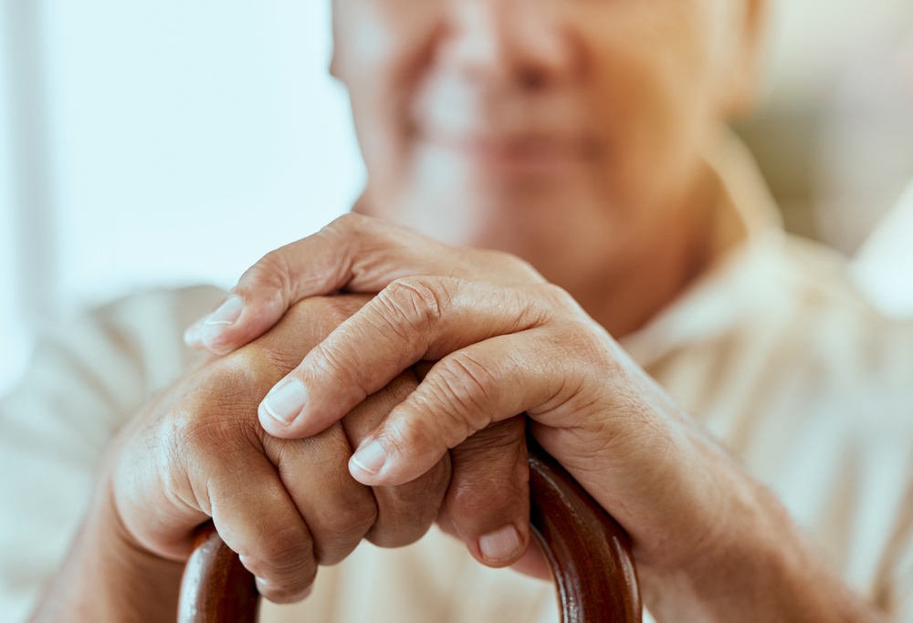 Fokus på två händer som håller i en käpp. I bakgrunden syns en ett ansikte av en äldre man. 