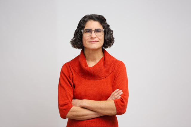 A person in a red sweater is standing against a white background. Their arms are crossed and they're looking into the camera.