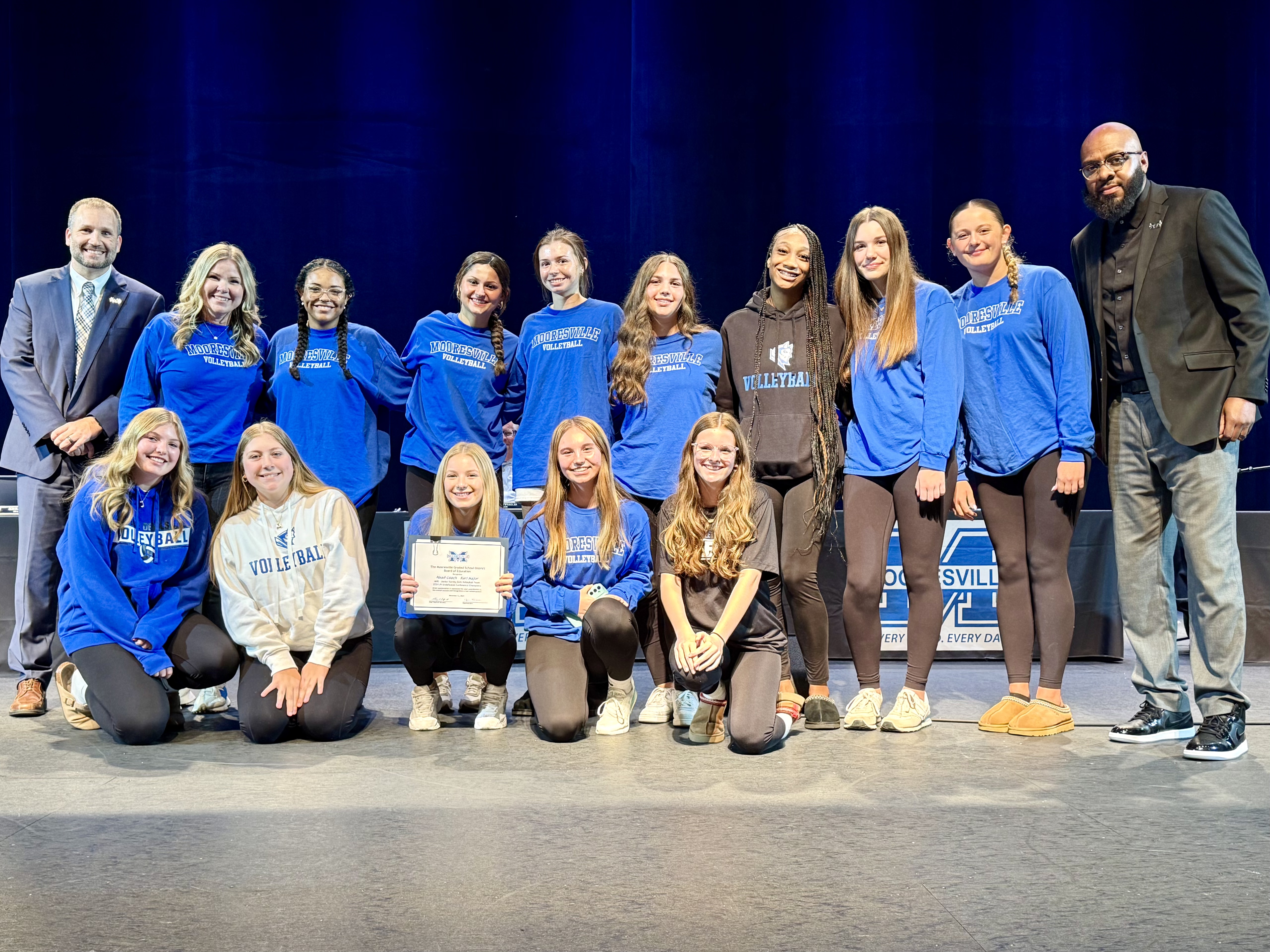 A group of female high school volleyball players stand between two male adults, along with their female adult coach.