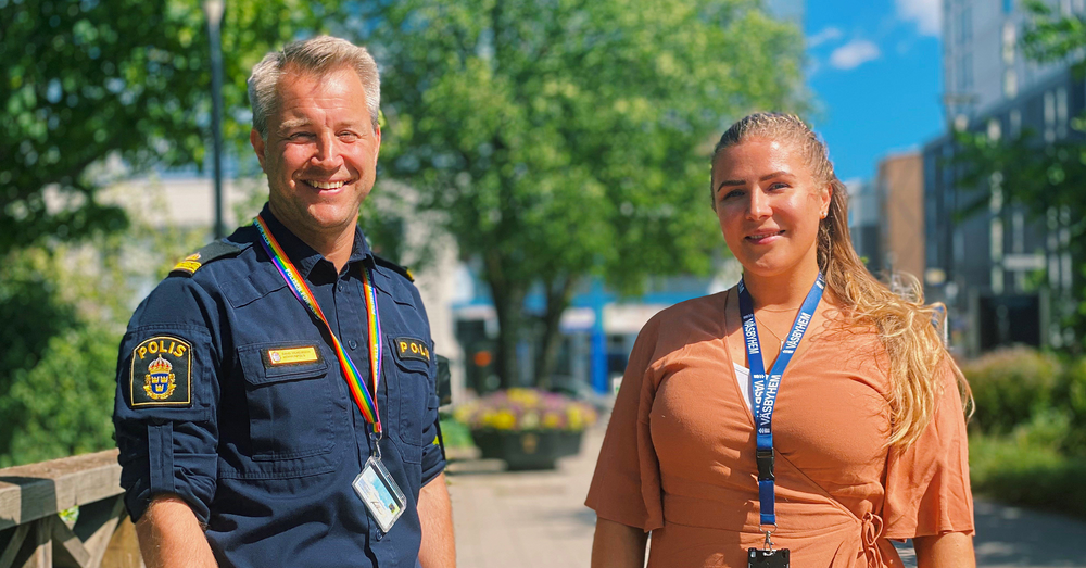 En man iklädd i polisuniform och en kvinna i orange klänning står intill ett stationsområde med mycket grönska i bakgrunden.