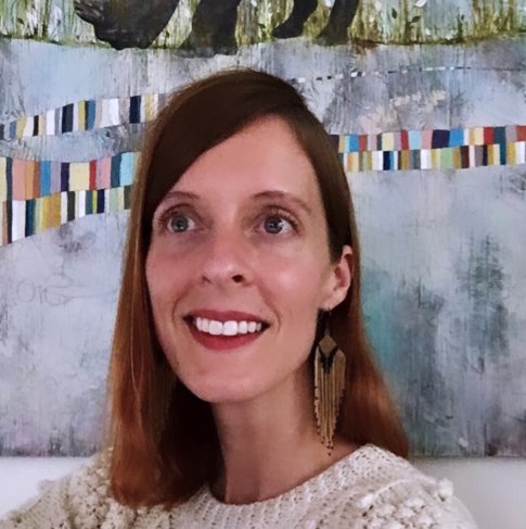 A person with red lipstick, a shoulder-length bob and dangly gold earrings is wearing a light colored sweater and smiling into the camera with a colorful background.