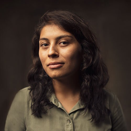 A person with long dark hair is wearing a green button-up shit and smiling at the camera with a dark background.