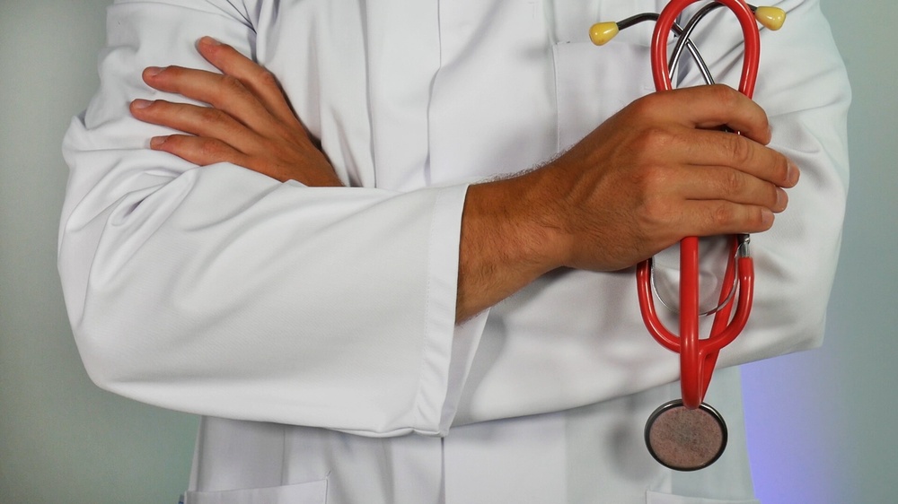 Bild på persons överkropp, står i vit rock, har armarna i kors och håller i ett par röda stetoskop.