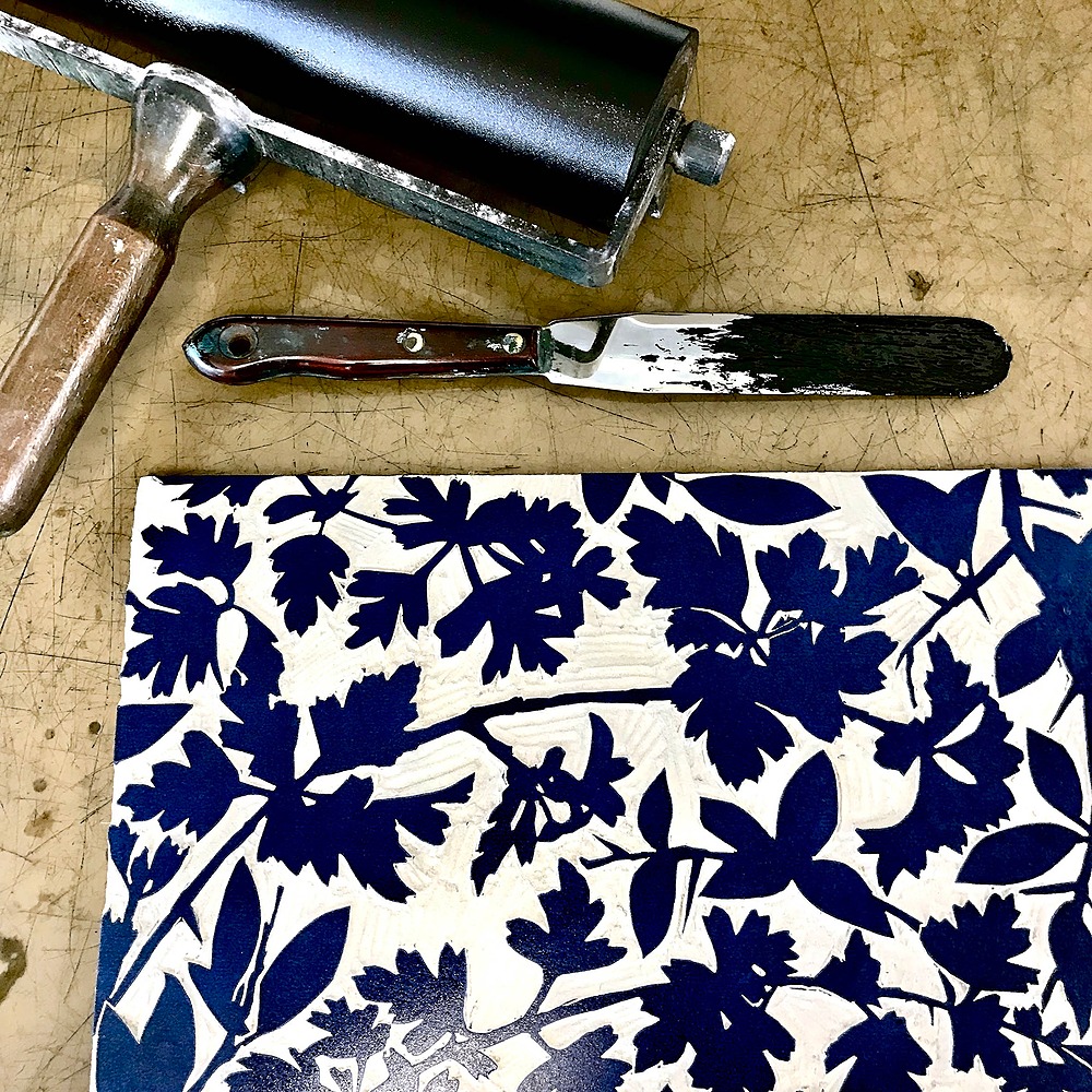 A lino block freshly inked and ready to be printed. The block features leafy foliage. Positioned with a brayer and ink knife next to the block.