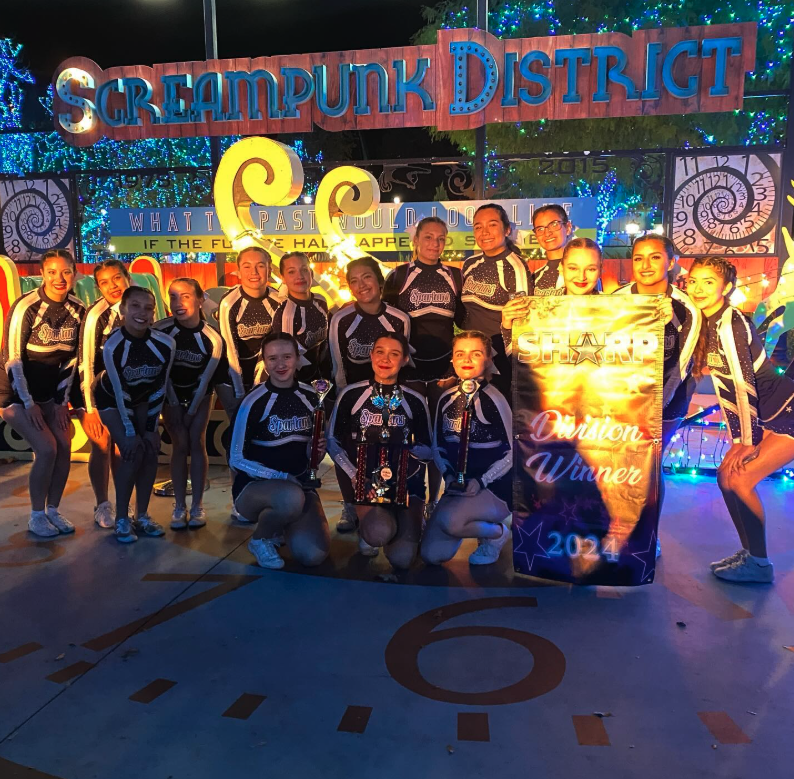 A cheerleading squad poses with trophies and a banner that reads 'Division Winner 2024' in front of a sign that says 'Screampunk District'.