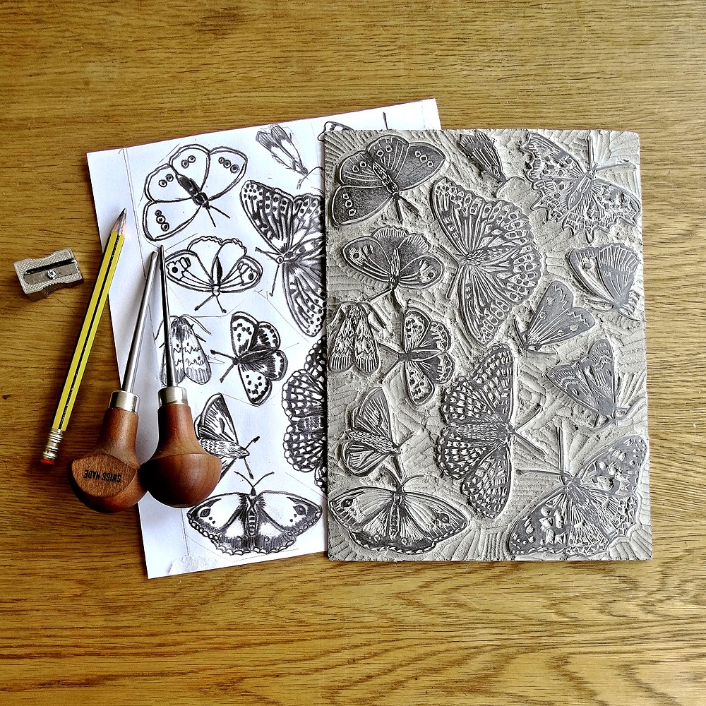 A carved lino block featuring moths is next to a piece of paper with the moth drawings used to transfer the design onto the block. On top of the piece of paper is a pencil sharpener, pencil and 2 carving tools.