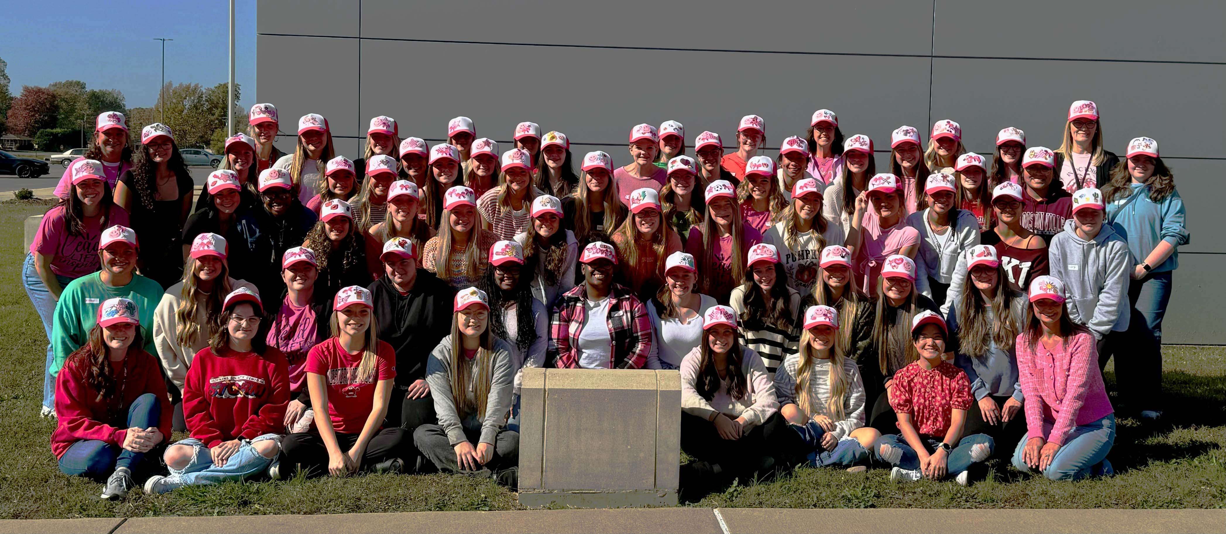 Group of about 50 people pose for photo outside wearing pink and white caps