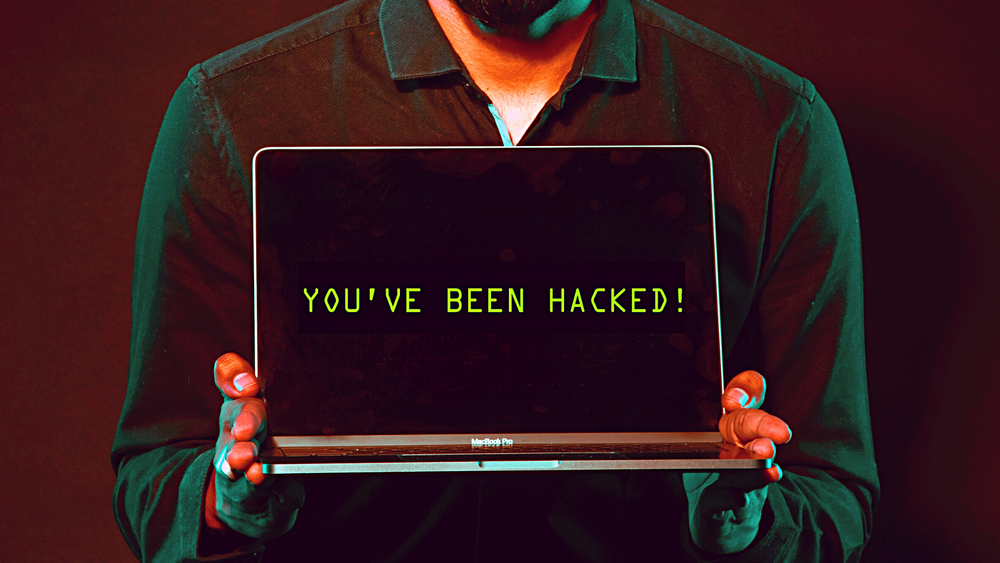 A man's upper body (face outside picture) in a dark shirt holding a laptop with a black screen and green text saying "YOU'VE BEEN HACKED".