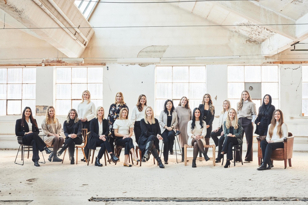 18 kvinnor står tillsammans i en stor fabrikslokal. Lokalen är ljus och kvinnorna står framför stora fönster. 