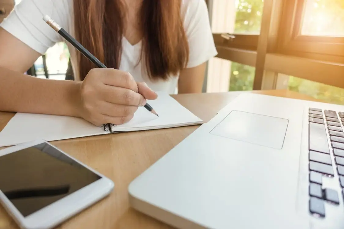 A woman writing on a piece of a paper in front of a laptop.