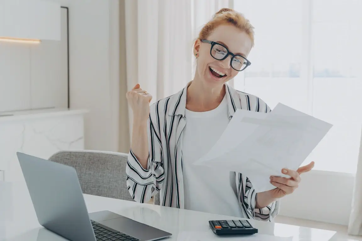 Overjoyed excited redhead woman in spectacles exclaiming yes with happy face expression as she holds papers with a calculator and laptop in front of her.