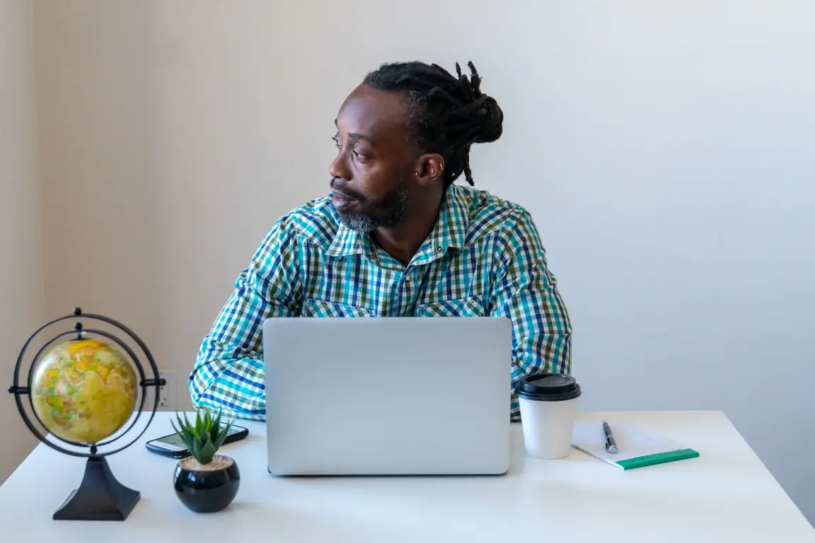 A photograph of a Black man sitting at his desk, looking out the window. On his desk, there's a small succulent and a mini globe. and