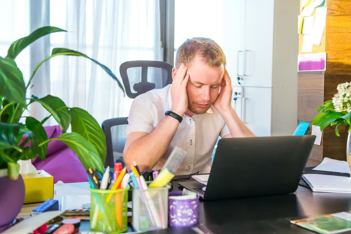 A man sitting at his desk in front of his laptop, rubbing his temples and feeling stressed.