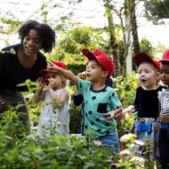 A photograph of a Black teacher working in education takes her Kindergarten students to a plant nursery to learn about ecology.